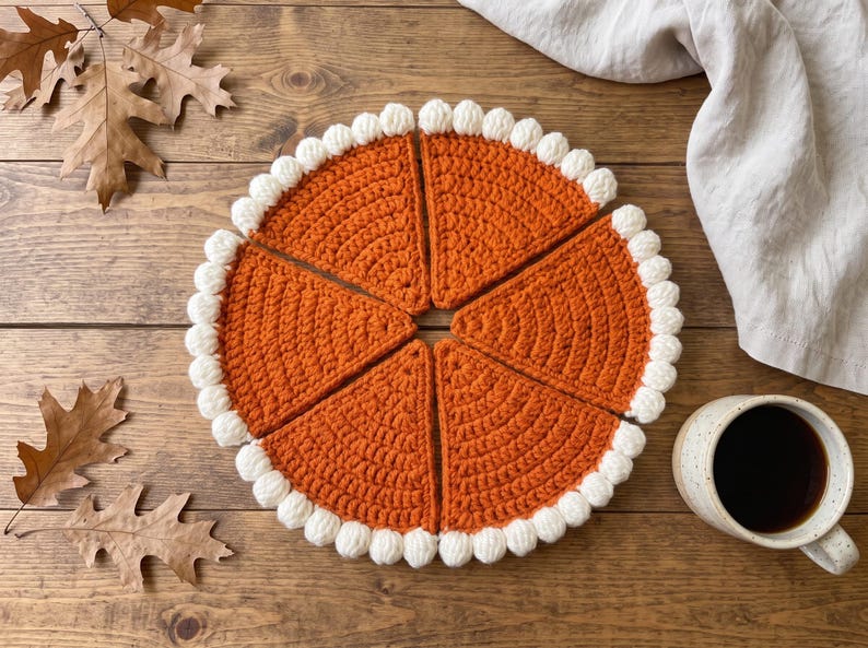 May include: A set of five triangular crochet pieces arranged in a circle to resemble a pumpkin pie. Each piece is made of orange yarn with a textured stitch pattern and features a border of white bobble stitches along the curved outer edge.