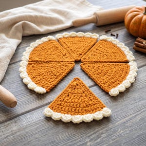 May include: Five triangular crochet pieces arranged to resemble a sliced pumpkin pie. Each piece features a textured orange center with a scalloped cream-colored border along the curved edge, mimicking a pie crust. The items are flat and handmade.