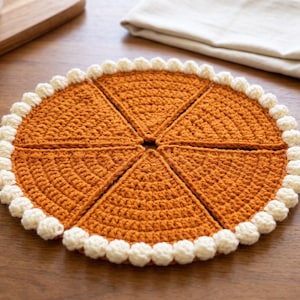 May include: A round, orange, crocheted trivet designed to resemble a sliced pie. The piece is composed of six triangular segments arranged in a circle, featuring a textured, cream-colored, bobble-stitch border around the entire outer edge.