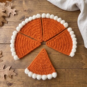 May include: Four triangular crochet pieces in a vibrant orange yarn, each featuring a textured white border of small, rounded bobble stitches along the curved edge, arranged to resemble individual slices of a pumpkin pie on a wooden surface.