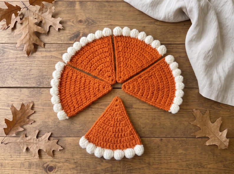 May include: Four triangular crochet pieces in a vibrant orange yarn, each featuring a textured white border of small, rounded bobble stitches along the curved edge, arranged to resemble individual slices of a pumpkin pie on a wooden surface.