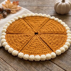 May include: A circular, golden-brown crocheted piece divided into six triangular segments, resembling a pumpkin pie. The edge is finished with a border of small, cream-colored crocheted bobbles, creating a textured, decorative circular mat.