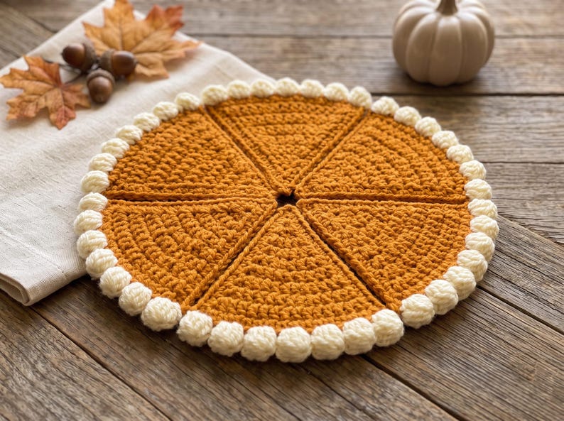 May include: A circular, golden-brown crocheted piece divided into six triangular segments, resembling a pumpkin pie. The edge is finished with a border of small, cream-colored crocheted bobbles, creating a textured, decorative circular mat.