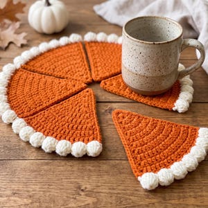 May include: A set of orange crochet coasters shaped like pie slices, each featuring a white bobble-stitch border along the curved edge. The coasters are arranged to form a circular pie shape, with one slice separated, and a speckled ceramic mug sits on top.