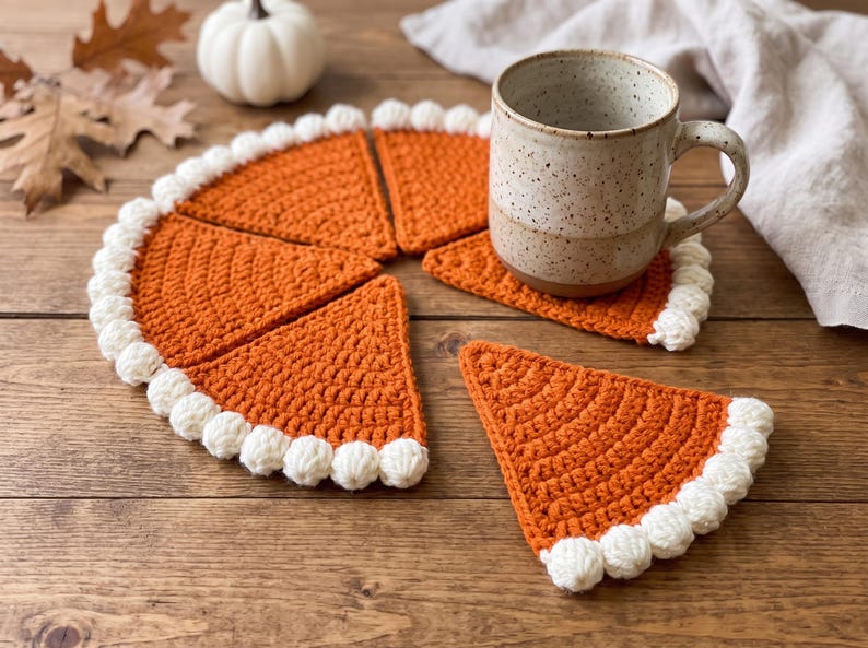 May include: A set of orange crochet coasters shaped like pie slices, each featuring a white bobble-stitch border along the curved edge. The coasters are arranged to form a circular pie shape, with one slice separated, and a speckled ceramic mug sits on top.
