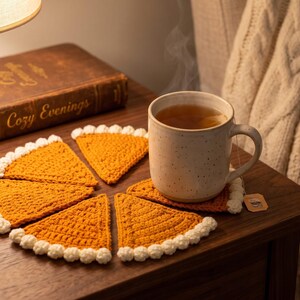 May include: A set of five triangular crochet coasters shaped like pumpkin pie slices, featuring a golden-orange textured center and a white scalloped border, arranged in a circular pattern around a speckled ceramic mug filled with hot tea.