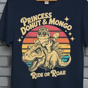 Puede incluir: Camiseta azul marino con un gráfico de un gato con gafas de sol montado en un dinosaurio, con el texto "PRINCESS DONUT & MONGO" y "RIDE OR ROAR". La camiseta tiene un diseño retro de puesta de sol.