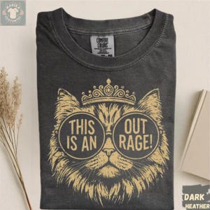 May include: Dark heather gray t-shirt featuring a cat wearing glasses and a crown. The cat graphic has the text "THIS IS AN OUT RAGE!". The shirt is made of a soft material and has a tag that says "Comfort Colors".