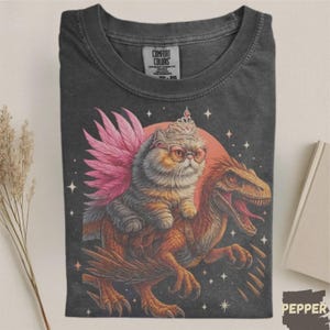 May include: Dark gray Comfort Colors t-shirt featuring a whimsical illustration. A cat wearing glasses and a crown with pink wings rides a dinosaur. The design includes a red sun and stars. The word "PEPPER" is in the bottom right corner.