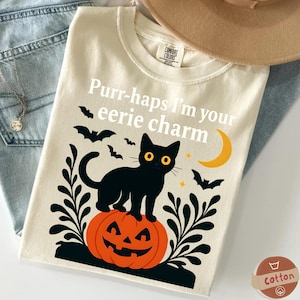 Pode incluir: Camiseta creme com um design de Halloween. O gráfico apresenta um gato preto com olhos amarelos em cima de uma abóbora laranja, rodeado por morcegos, uma lua crescente e o texto "Purr-haps I'm your eerie charm."