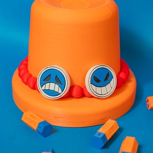 Peut inclure: Un chapeau orange avec deux motifs de visage circulaires bleus et blancs et un bandeau rouge perlé. Plusieurs objets hexagonaux orange et bleus sont dispersés autour du chapeau sur une surface bleue.