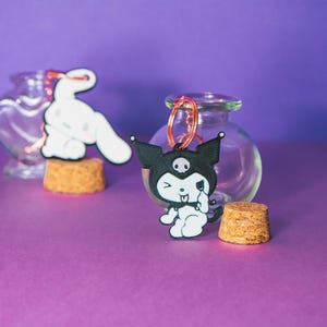 May include: A black and white Kuromi keyring with a copper-coloured ring, next to a clear glass bottle. A white cartoon character keyring is in the background. The items are on a purple surface.