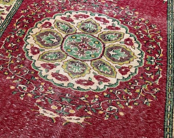 May include: A distressed, vintage rug with a deep red background and a central medallion design. The medallion features a floral pattern in shades of green, yellow, and cream. The rug's border is green and cream, adding to its antique aesthetic.