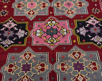 May include: A detailed, close-up view of a red, geometric patterned rug. The rug features repeating star and square shapes in shades of red, gray, black, pink, and yellow. The design is intricate, with detailed floral and geometric motifs throughout the rug's surface.