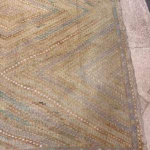 May include: A vintage rug with a geometric diamond pattern in shades of beige, blue, and brown. The rug has a fringed edge and a distressed appearance, suggesting age and use. The design features diagonal lines and dotted accents.