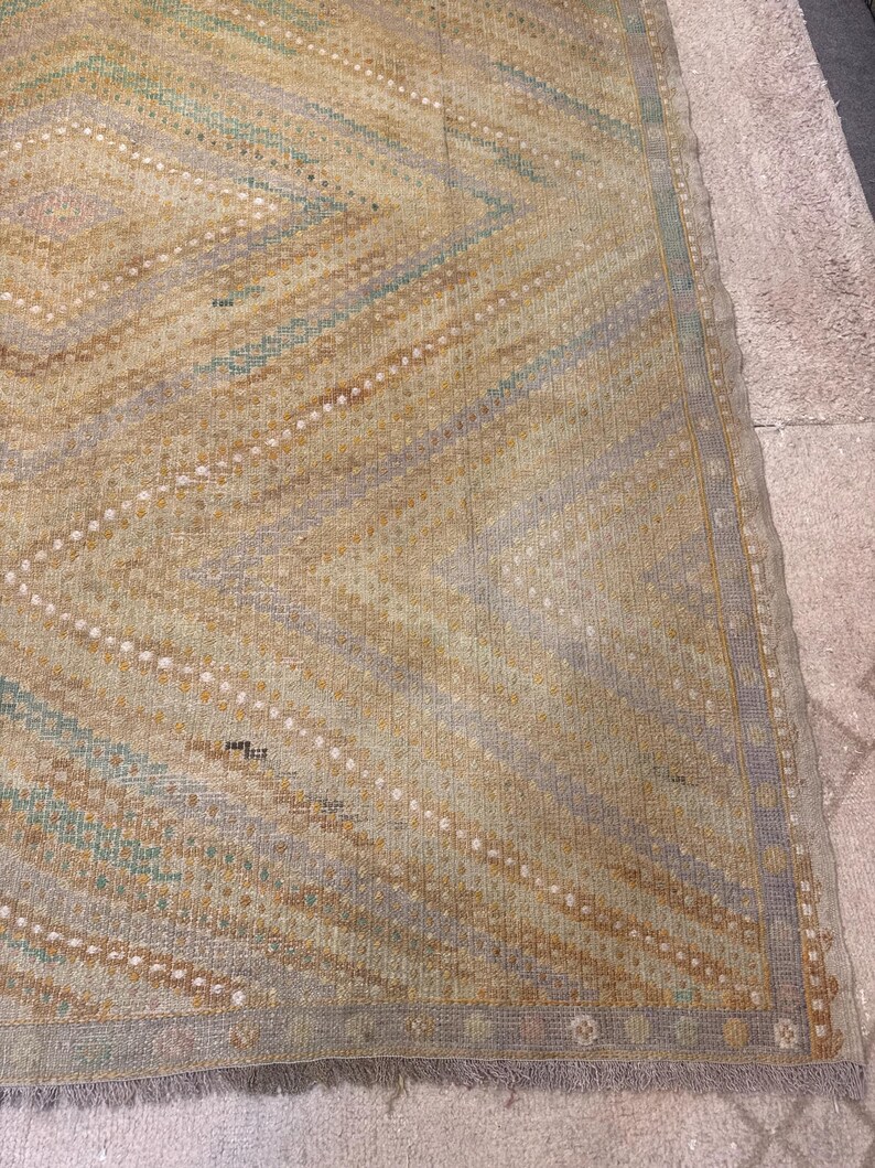 May include: A vintage rug with a geometric diamond pattern in shades of beige, blue, and brown. The rug has a fringed edge and a distressed appearance, suggesting age and use. The design features diagonal lines and dotted accents.