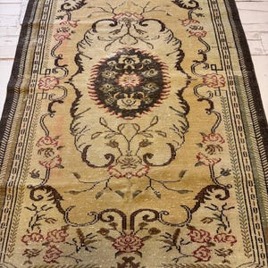 May include: An antique rug with a central medallion design. The rug features a beige background with floral and scroll patterns in shades of brown, red, and green. The border is a dark brown color.