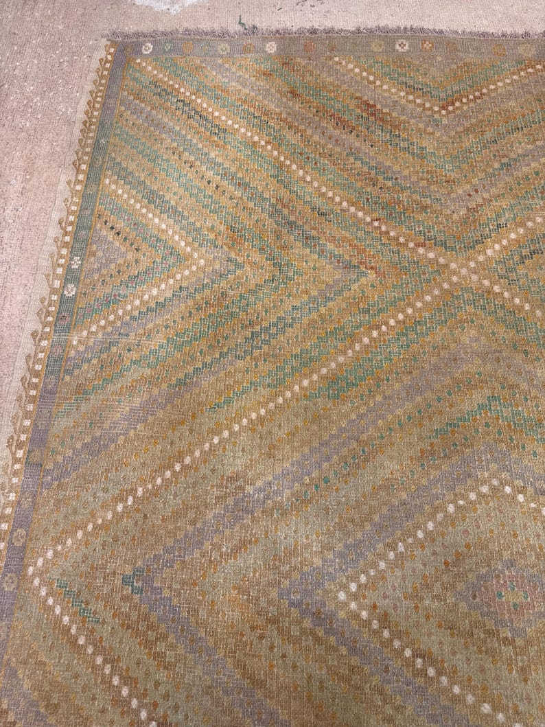 May include: A vintage rug with a geometric pattern. The rug features diagonal stripes in shades of green, gold, and lavender, with white dotted lines. The edges have a decorative border. The rug is likely made of woven material.