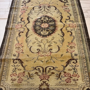 May include: A rectangular, vintage rug with a central medallion design. The rug features a beige background with floral and scroll patterns in shades of brown, red, and green. The border is a dark brown with a repeating geometric pattern. The rug is likely made of wool.