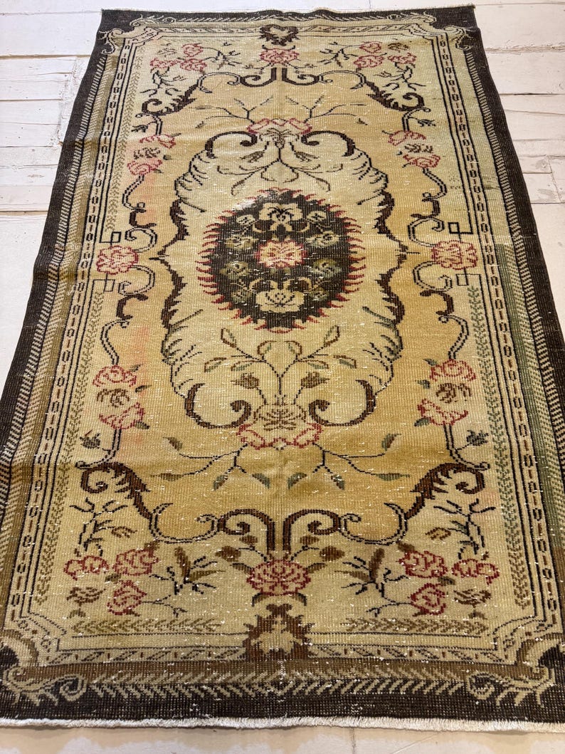 May include: A rectangular, vintage rug with a central medallion design. The rug features a beige background with floral and scroll patterns in shades of brown, red, and green. The border is a dark brown with a repeating geometric pattern. The rug is likely made of wool.