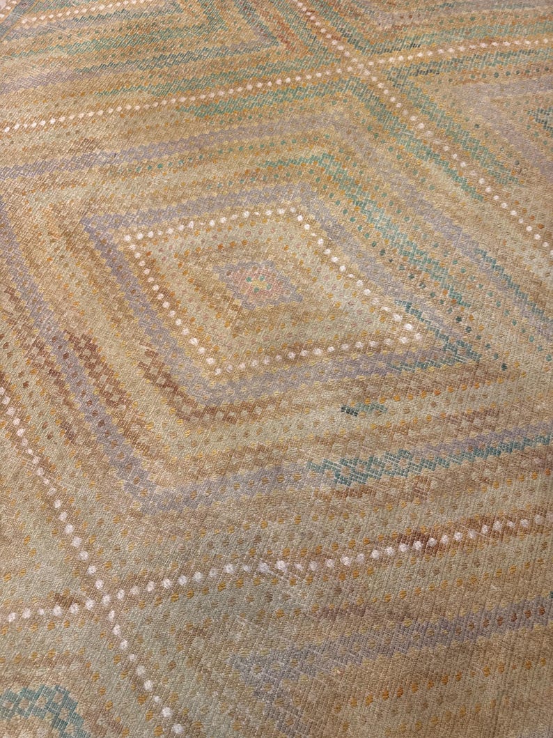 May include: Close-up of a woven textile with a geometric diamond pattern. The design features alternating diagonal stripes in shades of tan, green, and lavender, with rows of white dots. The overall texture appears soft and slightly worn.