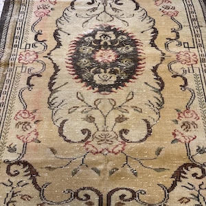 May include: A rectangular, vintage rug with an intricate floral design. The rug features a cream-colored background with a central medallion and floral motifs in shades of brown, red, and green. The border is a darker brown with geometric patterns.