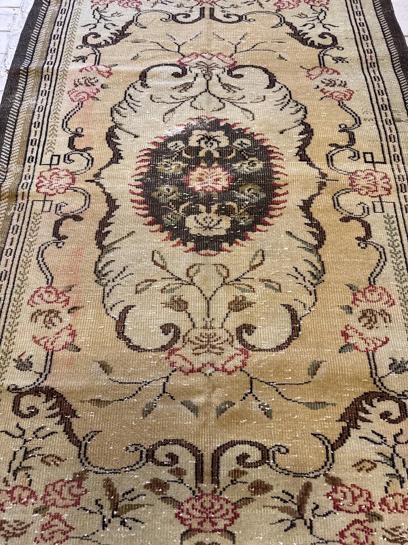 May include: A rectangular, vintage rug with an intricate floral design. The rug features a cream-colored background with a central medallion and floral motifs in shades of brown, red, and green. The border is a darker brown with geometric patterns.