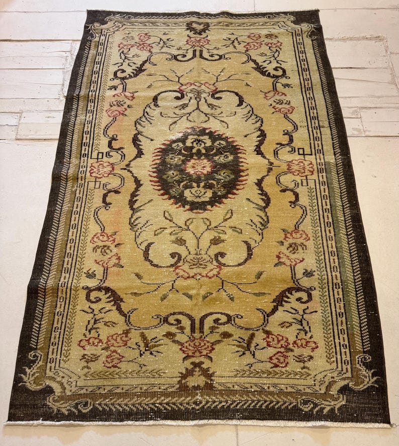 May include: Rectangular vintage rug with a central medallion and floral motifs. The rug features a beige field with a dark brown border and accents in red and green. The design includes stylized flowers and intricate patterns, suitable for home decor.