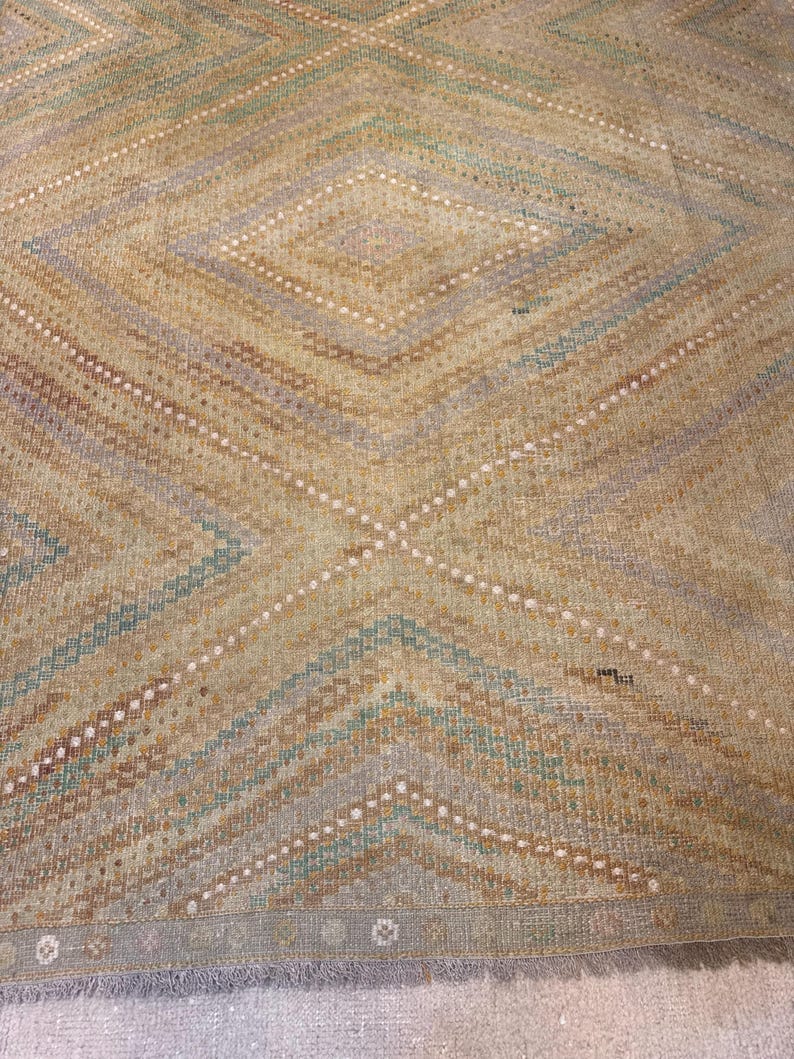May include: A vintage rug with a geometric diamond pattern. The rug features a color palette of beige, light blue, and brown, with white dotted lines accenting the design. The edges have a fringe detail.