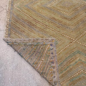 May include: A vintage rug with a geometric diamond pattern in shades of green, tan, and gray. The rug has a distressed look with visible wear and tear, and a fringed edge. The corner of the rug is folded over, revealing the underside.