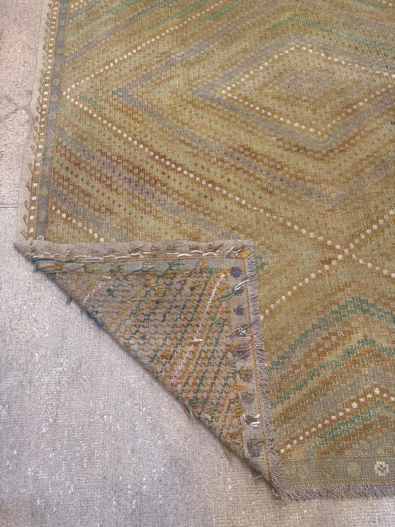 May include: A vintage rug with a geometric diamond pattern in shades of green, tan, and gray. The rug has a distressed look with visible wear and tear, and a fringed edge. The corner of the rug is folded over, revealing the underside.