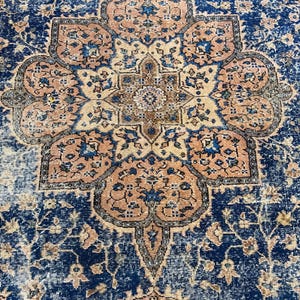 May include: A detailed close-up of a vintage rug with a central medallion design. The rug features a navy blue background with a faded, distressed appearance. The medallion is a combination of peach, beige, and blue floral patterns, creating a classic, ornate aesthetic.