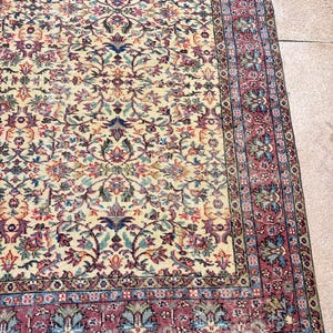May include: A detailed shot of a vintage rug with a cream-colored central field and an intricate floral pattern in shades of red, blue, and green. The rug has a burgundy border with a repeating design, and a fringed edge.