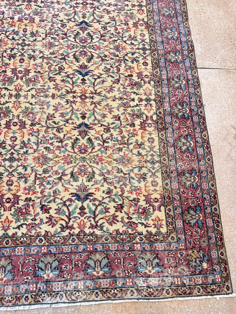 May include: A detailed shot of a vintage rug with a cream-colored central field and an intricate floral pattern in shades of red, blue, and green. The rug has a burgundy border with a repeating design, and a fringed edge.