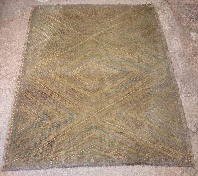 May include: A rectangular rug with a geometric diamond pattern. The rug features a color palette of muted greens, browns, and beige. The design includes intersecting lines and diamond shapes, creating a textured appearance. The rug has a fringe border.