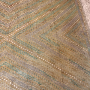 May include: A close-up of a vintage rug with a geometric pattern. The rug features a chevron design in shades of beige, gold, teal, and lavender. White dotted lines accent the diagonal stripes. The rug has a fringed edge.