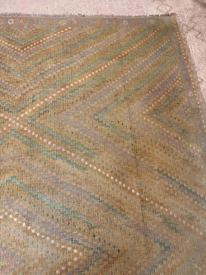 May include: A close-up of a vintage rug with a geometric pattern. The rug features a chevron design in shades of beige, gold, teal, and lavender. White dotted lines accent the diagonal stripes. The rug has a fringed edge.