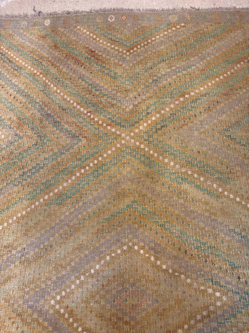 May include: A close-up of a woven rug with a geometric pattern. The rug features a repeating design of diagonal lines in shades of tan, green, and blue, with white dotted accents. The rug has a fringed edge.