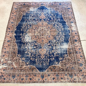 May include: An antique Persian rug with a central medallion design. The rug features a deep blue field with a distressed appearance, framed by a border in shades of brown and pink. Floral and geometric patterns are visible throughout the rug's design.