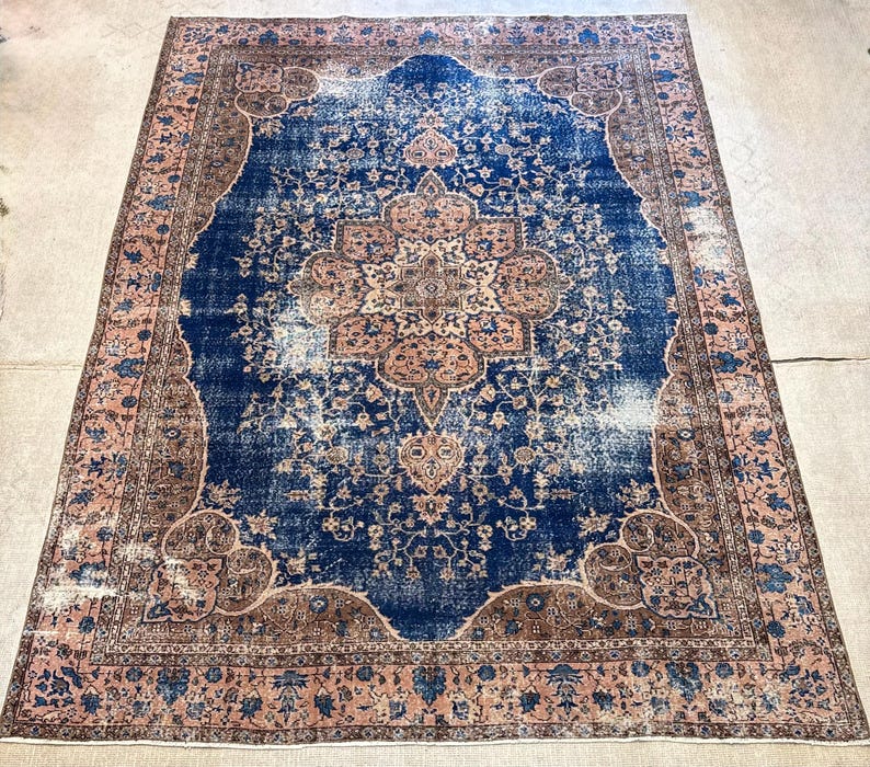 May include: An antique Persian rug with a central medallion design. The rug features a deep blue field with a distressed appearance, framed by a border in shades of brown and pink. Floral and geometric patterns are visible throughout the rug's design.