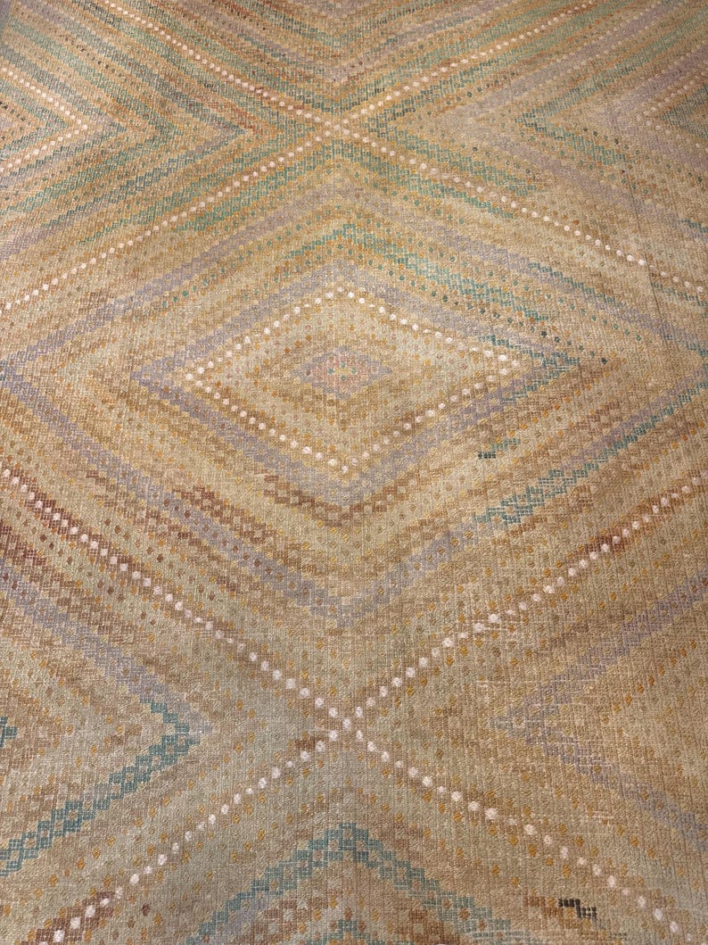 May include: Close-up of a woven rug with a diamond pattern. The rug features a beige background with diamond shapes in shades of brown, blue, and purple. White dotted lines accent the diamond shapes. The rug has a textured appearance.
