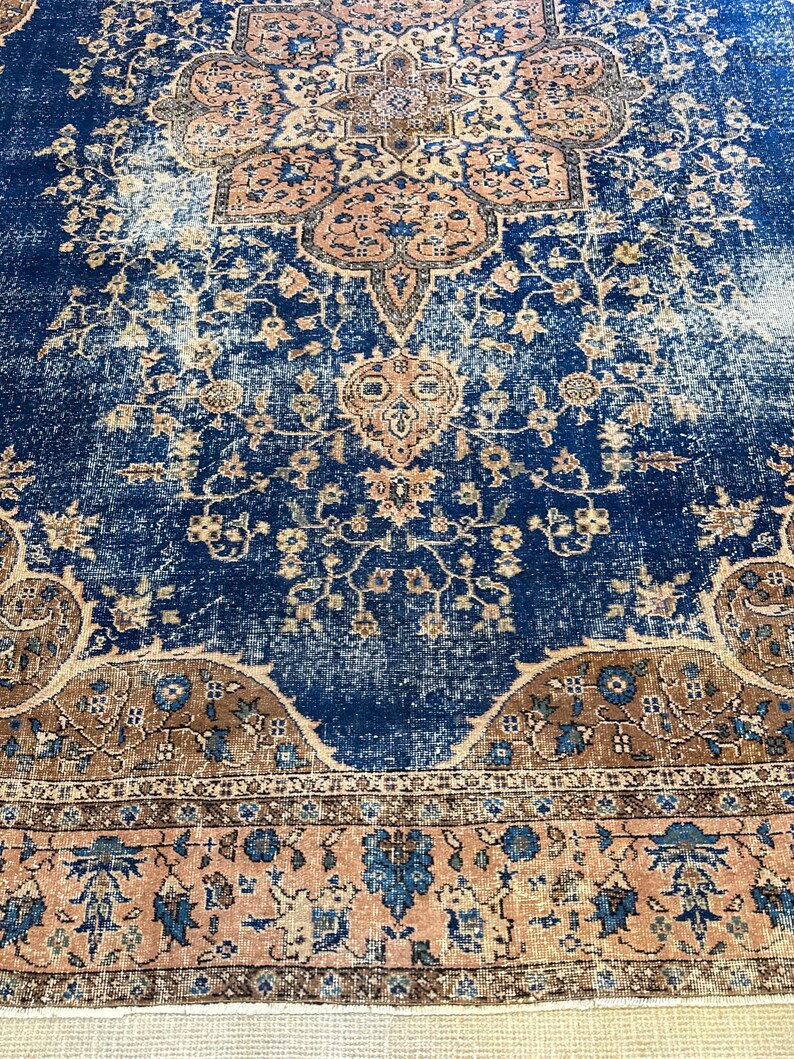 May include: A distressed, vintage-style rug with a rich navy blue central field. The rug features intricate floral and geometric patterns in shades of peach, brown, and cream. The design includes a detailed border.