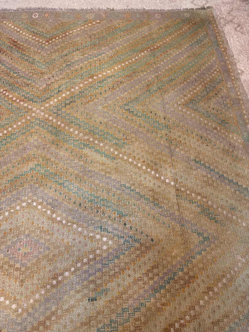 May include: A vintage rug with a geometric pattern. The rug features a beige background with diagonal stripes in shades of brown, green, and purple. White dots accent the design. The rug has a fringed edge.