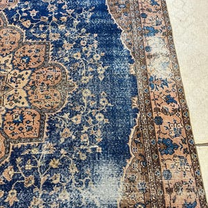 May include: A distressed, vintage-style rug with a central field of deep blue, adorned with floral and geometric patterns in shades of peach, brown, and cream. The rug's edges feature a detailed border, adding to its antique aesthetic.