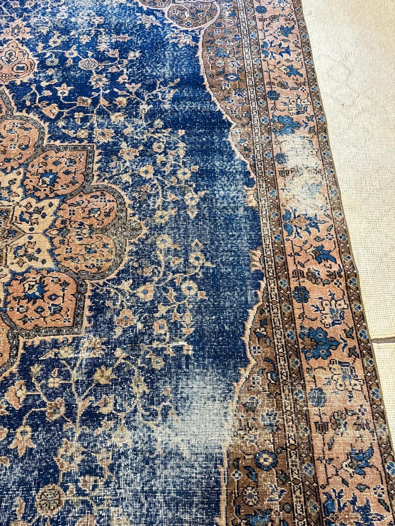 May include: A distressed, vintage-style rug with a central field of deep blue, adorned with floral and geometric patterns in shades of peach, brown, and cream. The rug's edges feature a detailed border, adding to its antique aesthetic.