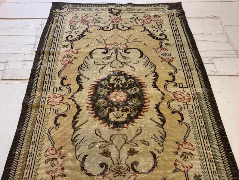 May include: An antique beige rug with a central medallion design and floral motifs. The rug features a dark brown border and accents in red and black. The intricate pattern and faded appearance suggest a vintage origin, adding a touch of elegance to any space.