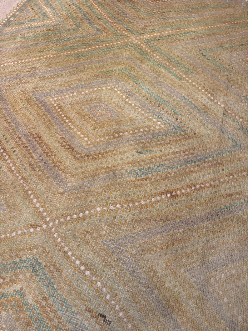 May include: A close-up of a rug with a geometric diamond pattern. The rug features a color palette of tan, brown, and blue-green hues. The design includes diagonal lines and diamond shapes, creating a textured appearance. The rug's construction appears to be woven.