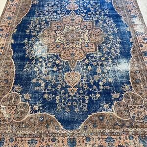 May include: A large, rectangular rug with a distressed, vintage appearance. The rug features a central medallion design in shades of tan and brown, set against a deep blue field. The border is a mix of tan, blue, and brown floral patterns.