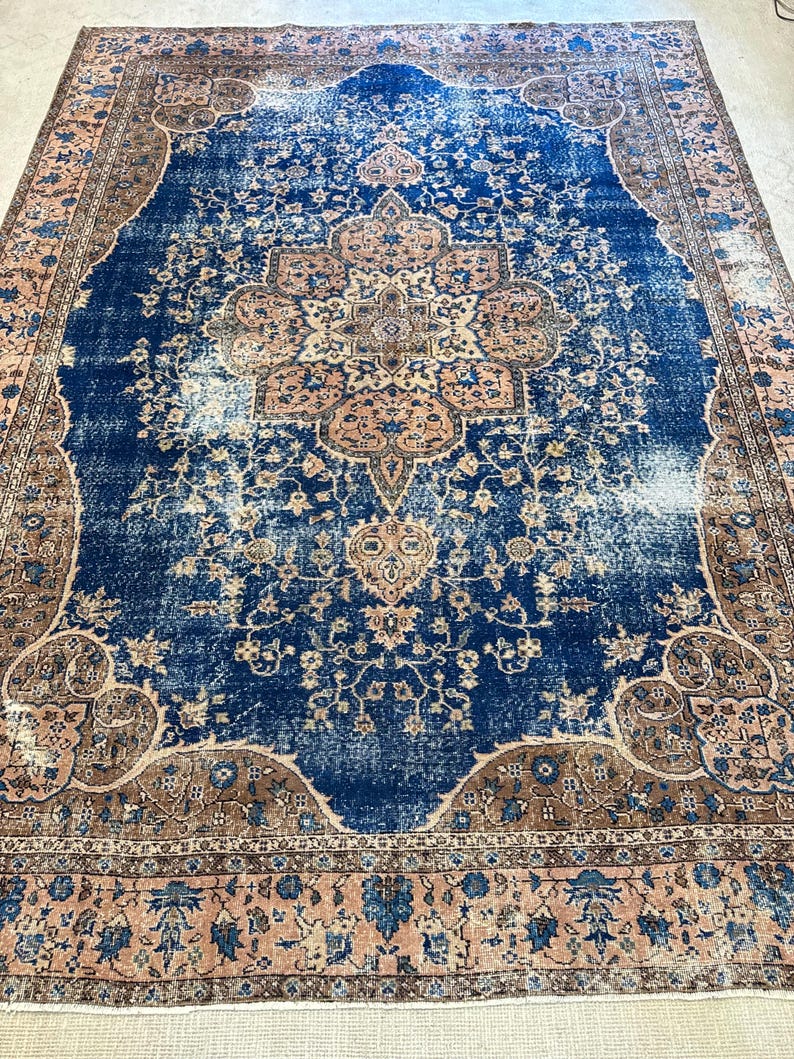 May include: A large, rectangular rug with a distressed, vintage appearance. The rug features a central medallion design in shades of tan and brown, set against a deep blue field. The border is a mix of tan, blue, and brown floral patterns.