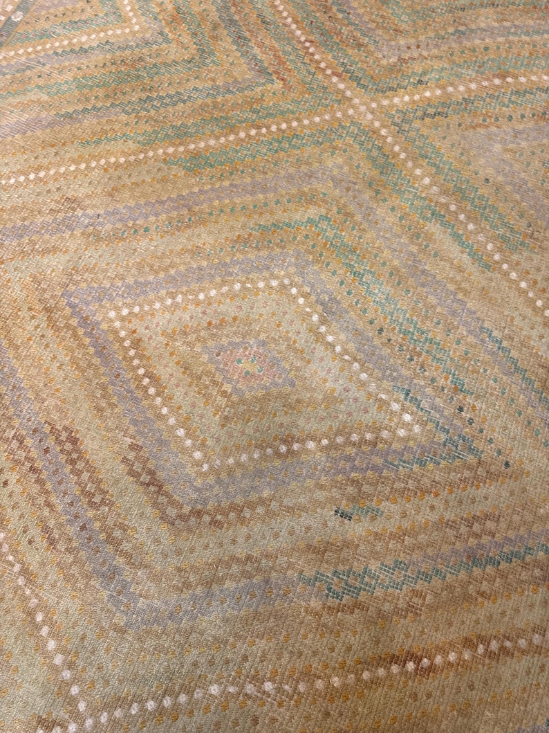 May include: Close-up of a woven textile with a geometric diamond pattern. The design features alternating bands of tan, light blue, and lavender hues, accented with rows of white dots. The overall texture appears soft and inviting.